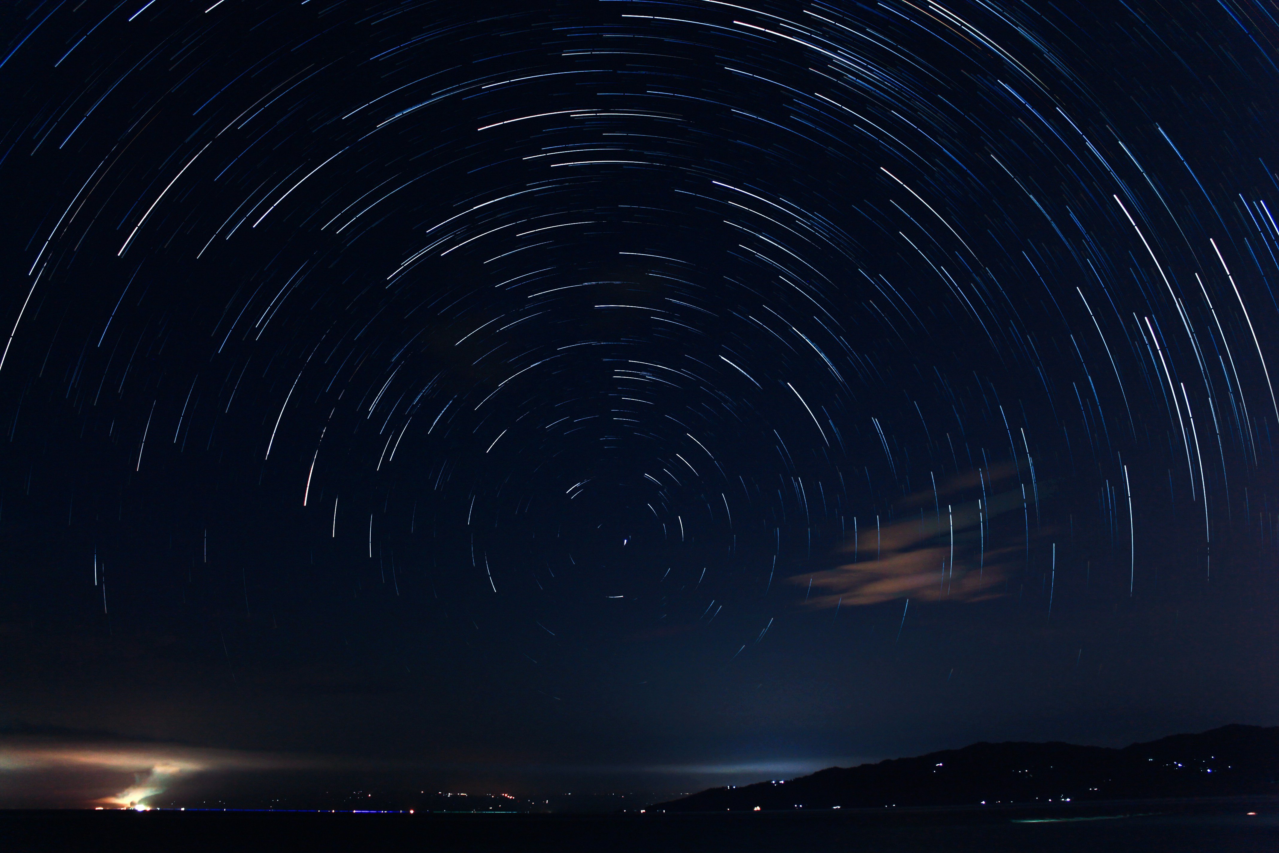 Sternspuren am Nachthimmel über einer dunklen Landschaft mit lichtern Ortschaften am Horizont.